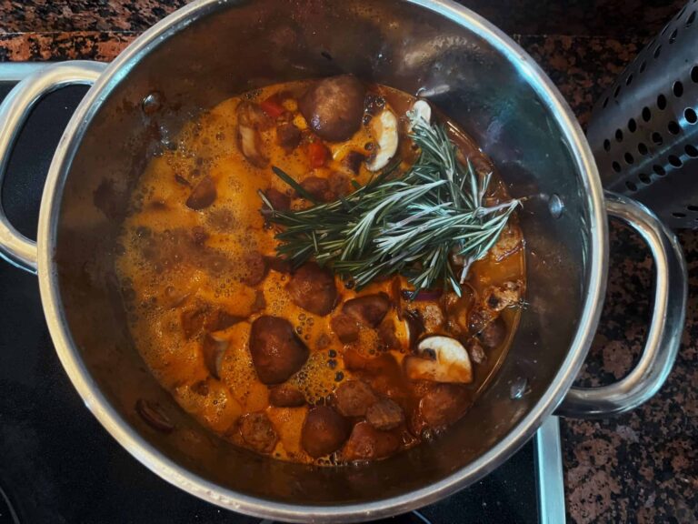 Veganes Gulasch mit Sojaschnetzeln, Pilzen und einem Zweig Rosmarin köchelt im Topf.